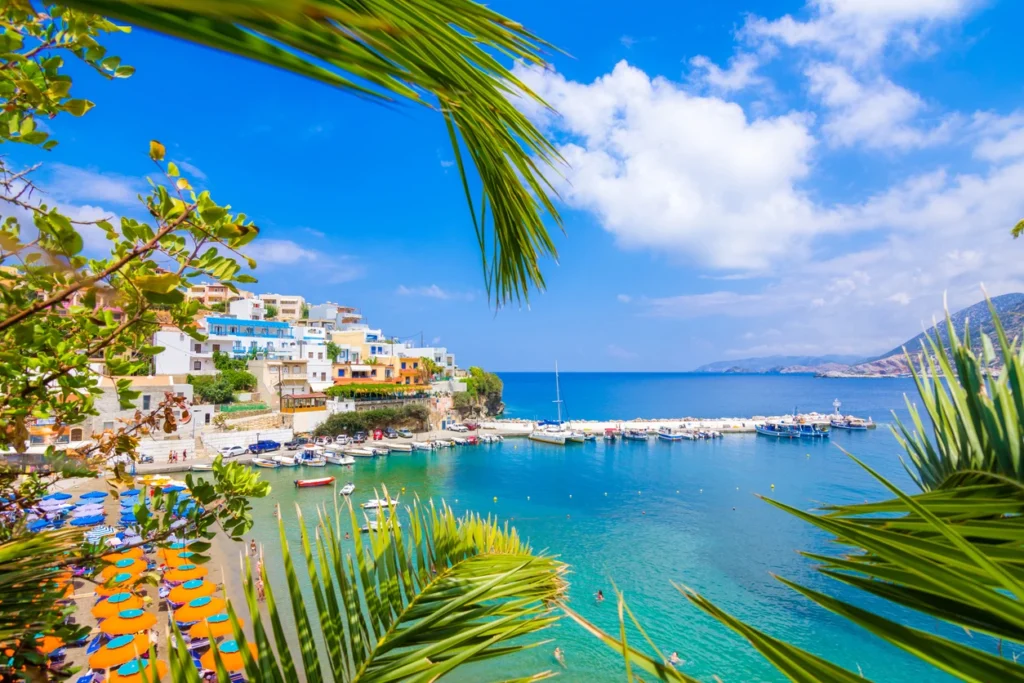 A vibrant view of the Old Venetian Harbor in Rethymno, Crete, framed by green palm branches. The scene shows the waterfront with its traditional colorful buildings and a lighthouse, with small fishing boats docked in the turquoise water under a blue sky.