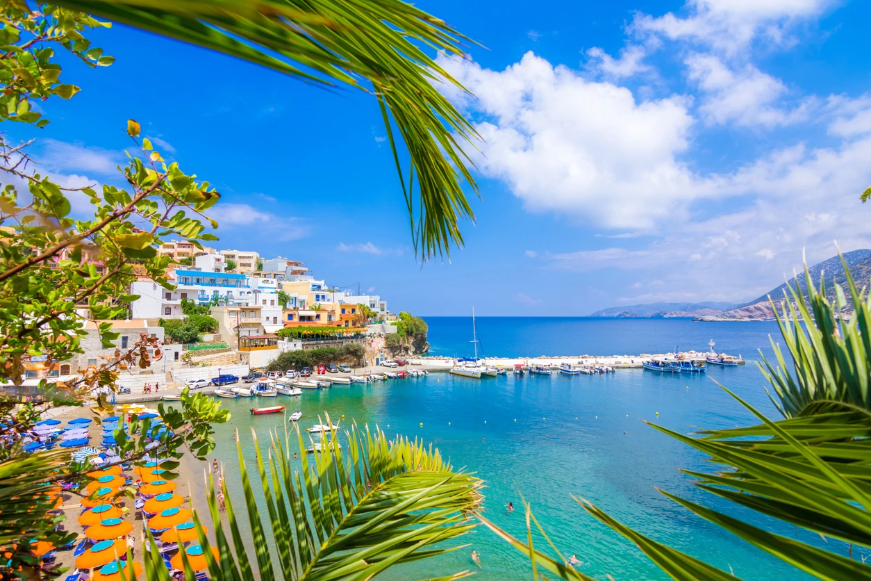 A vibrant view of the Old Venetian Harbor in Rethymno, Crete, framed by green palm branches. The scene shows the waterfront with its traditional colorful buildings and a lighthouse, with small fishing boats docked in the turquoise water under a blue sky.