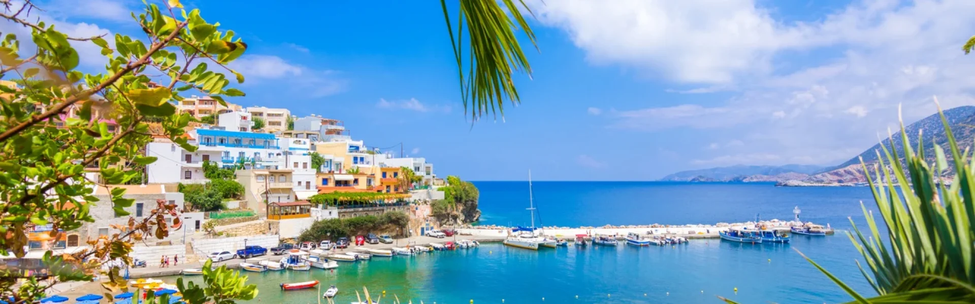 A vibrant view of the Old Venetian Harbor in Rethymno, Crete, framed by green palm branches. The scene shows the waterfront with its traditional colorful buildings and a lighthouse, with small fishing boats docked in the turquoise water under a blue sky.